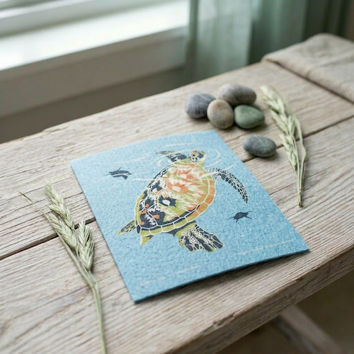 Blue Swedish dishcloth with a sea turtle print, on a wooden table with stones and dried wheat.