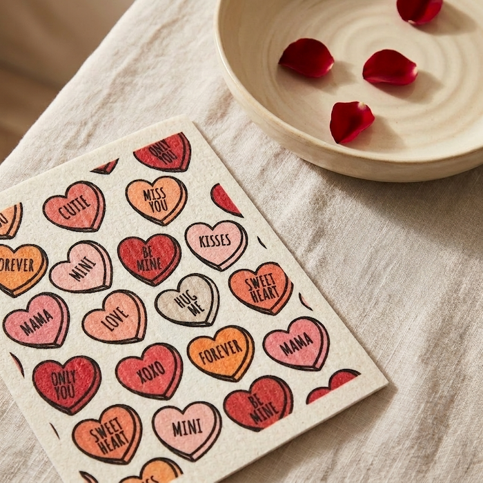 Driftless Studio Swedish dishcloth with red, pink, and orange candy heart pattern on a linen tablecloth next to a bowl with