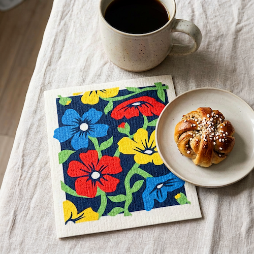 Swedish dishcloth in Flower Festival pattern featuring colorful flowers on a dark blue background with a cup of coffee and